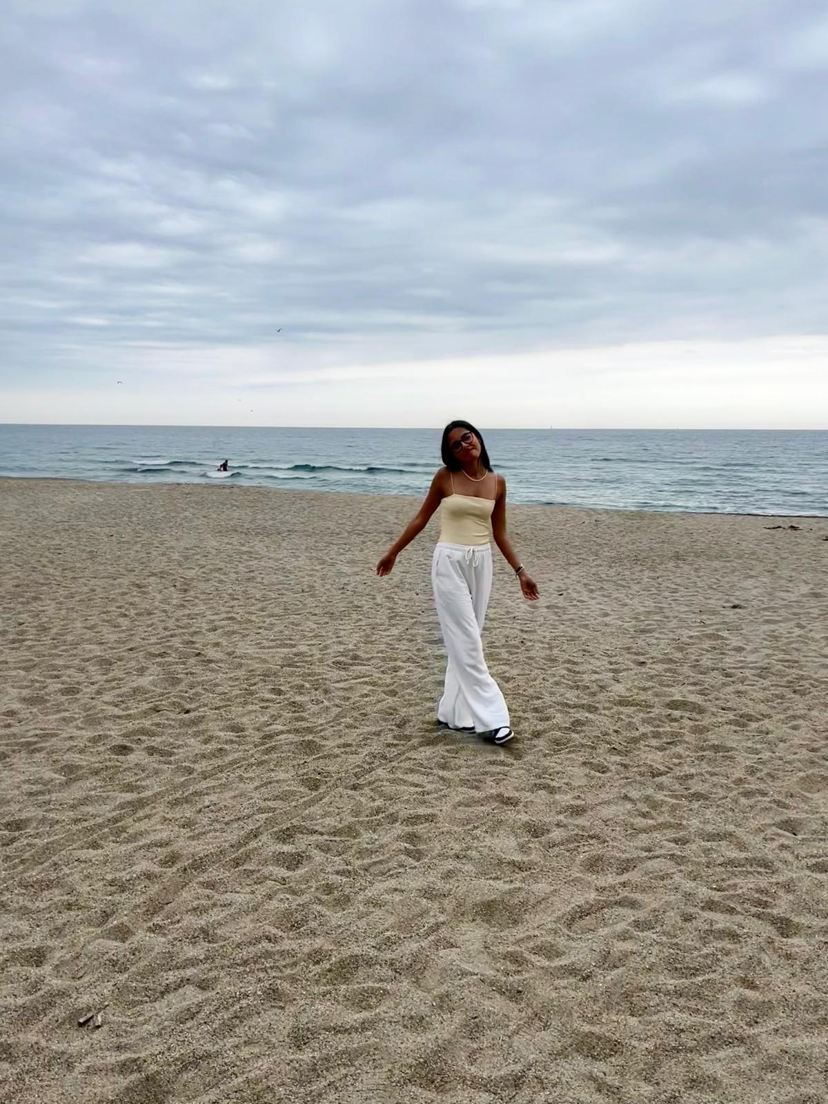 Photo of woman with tanned skin and brown hair on the beach. She is wearing white trousers and a yellow top