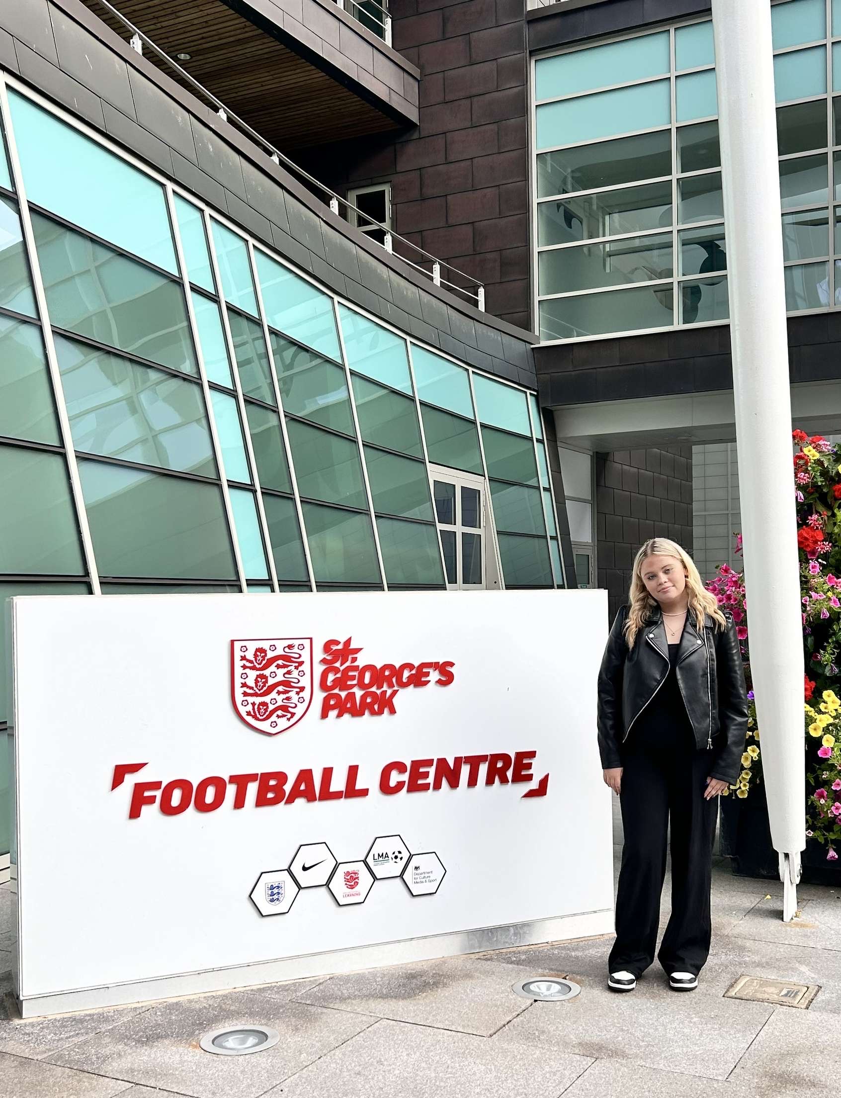 Photo of a white woman with blonde hair wearing all black standing in front of St George's Park sign