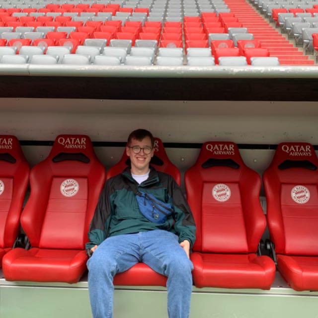 Photo of a white man with brown hair and glasses sitting in the dug out at a football stadium