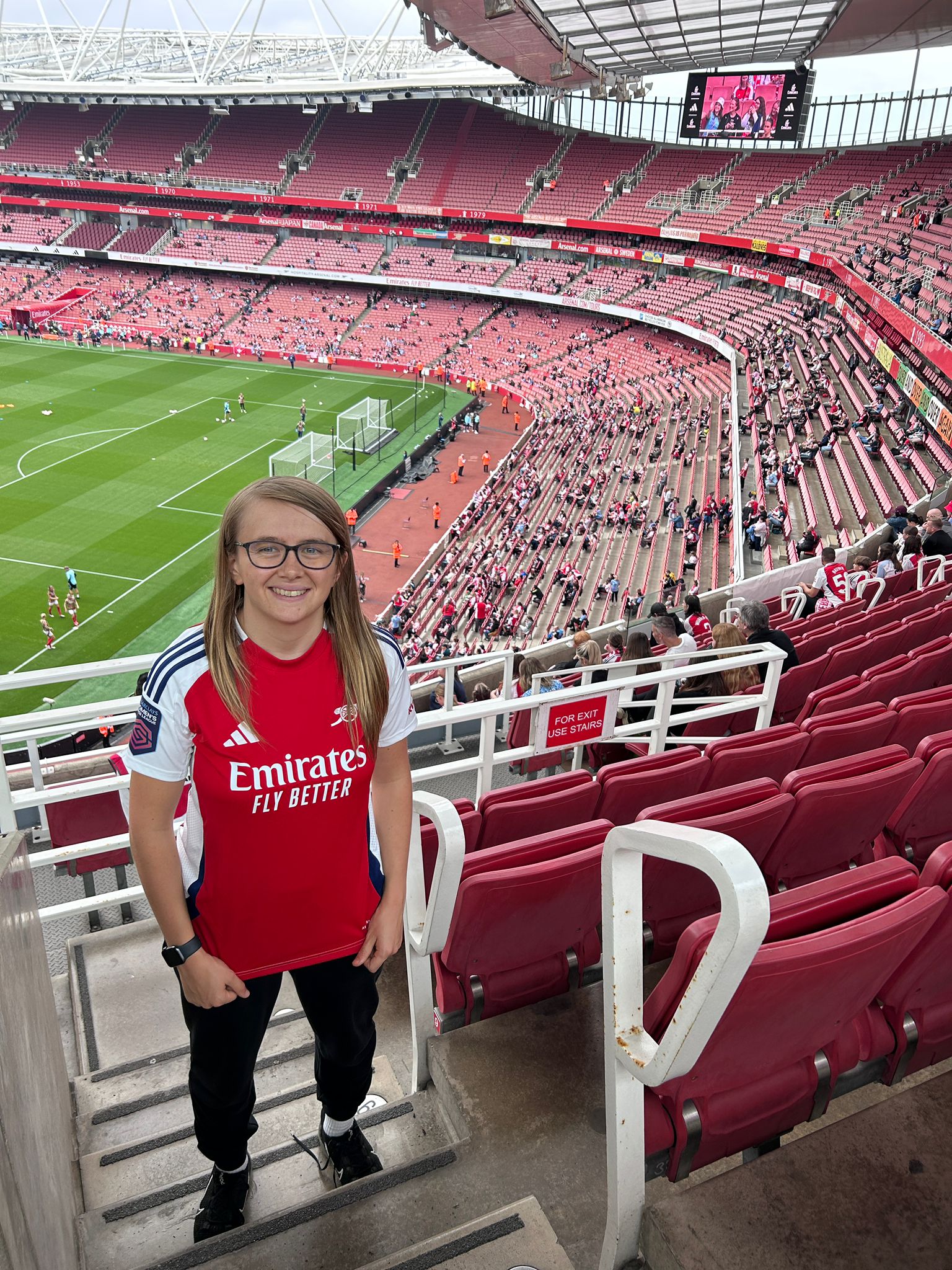 Photo of a white woman with glasses standing at the Emirates stadium wearing an arsenal top