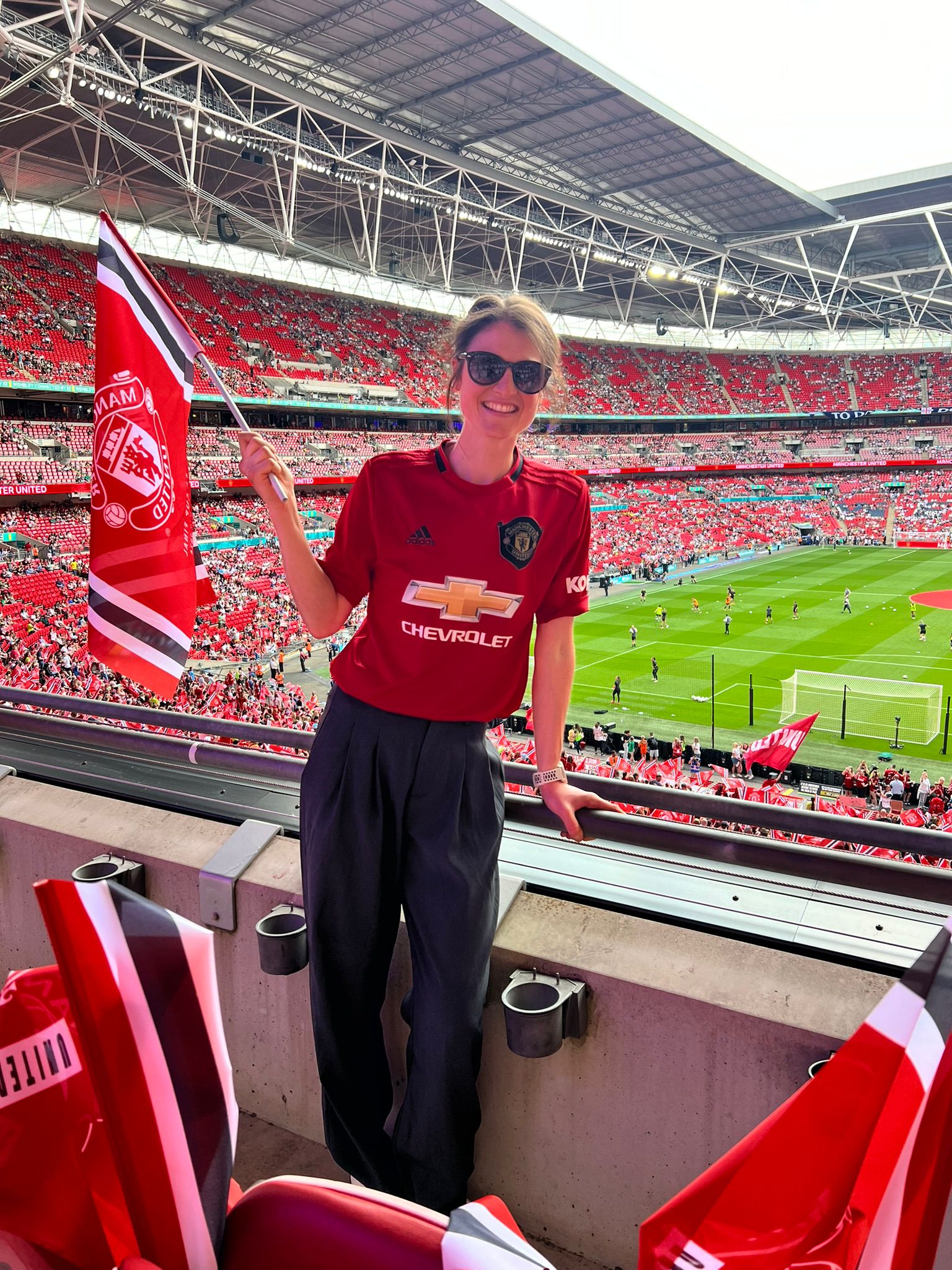 Photo of white woman wearing Manchester United red shirt, holding flag and standing in Old Trafford stadium.