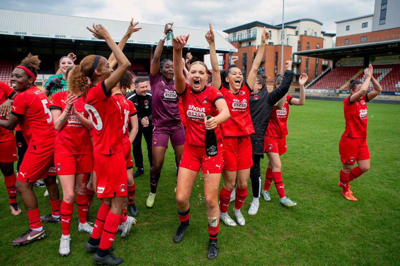 Leyton Orient Women celebrating in red kits, cheering and smiling with champagne and jumping up and down.