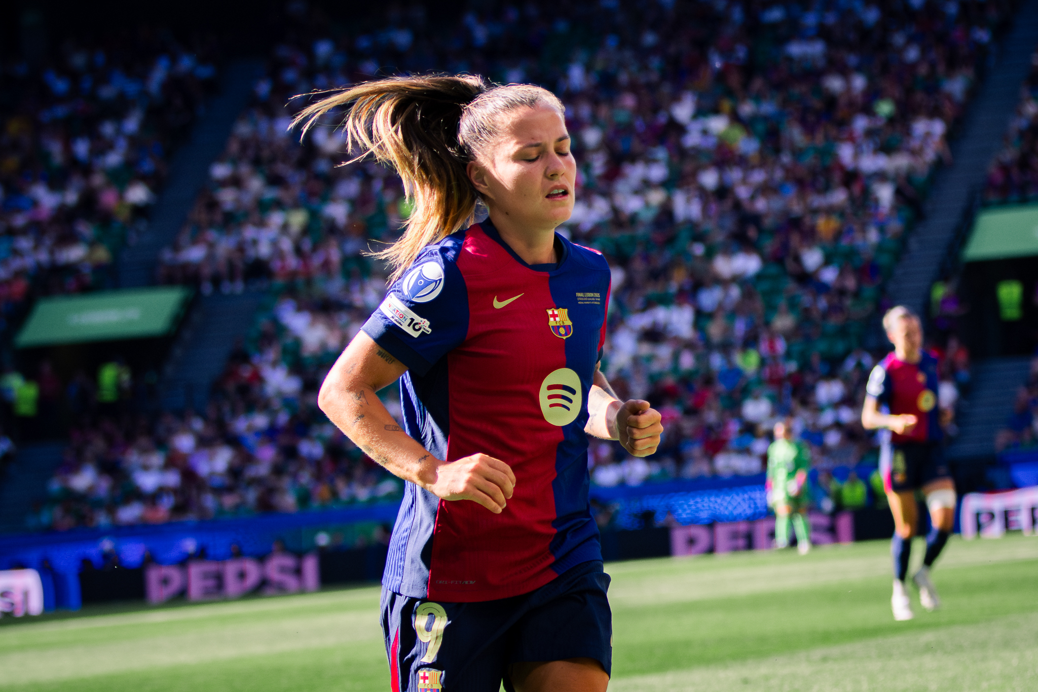 A female FC Barcelona player, Claudia Pina, in a red and blue jersey with the number 9 is running on the field during a match. Her eyes are closed and her ponytail is swinging as she moves. The stadium is full of spectators, and other players are visible in the background. The scene is brightly lit with sunlight.