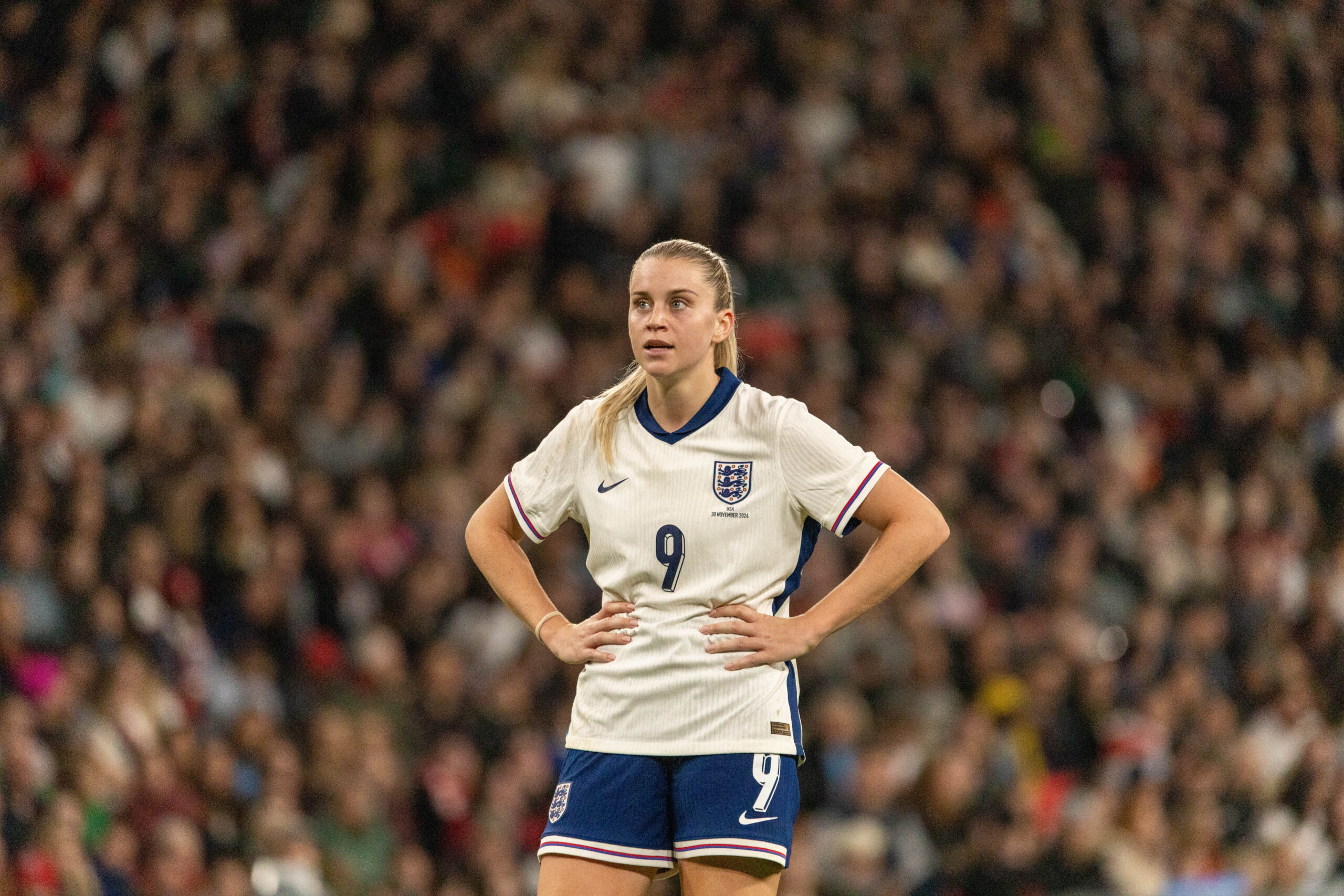 Alessia Russo in white jersey number 9 standing on the field with hands on hips, looking focused during a match, with a blurred crowd in the background.