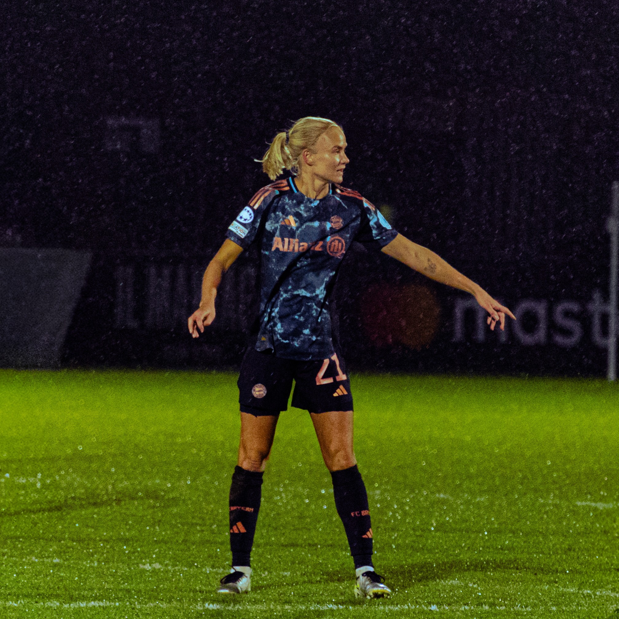 Pernille Harder wearing a dark camouflage-patterned Bayern Munich away kit with the number 21 is standing on a wet, rain-soaked pitch at night. She has blonde hair tied in a ponytail and is pointing with her right arm, appearing to communicate with teammates during a match.