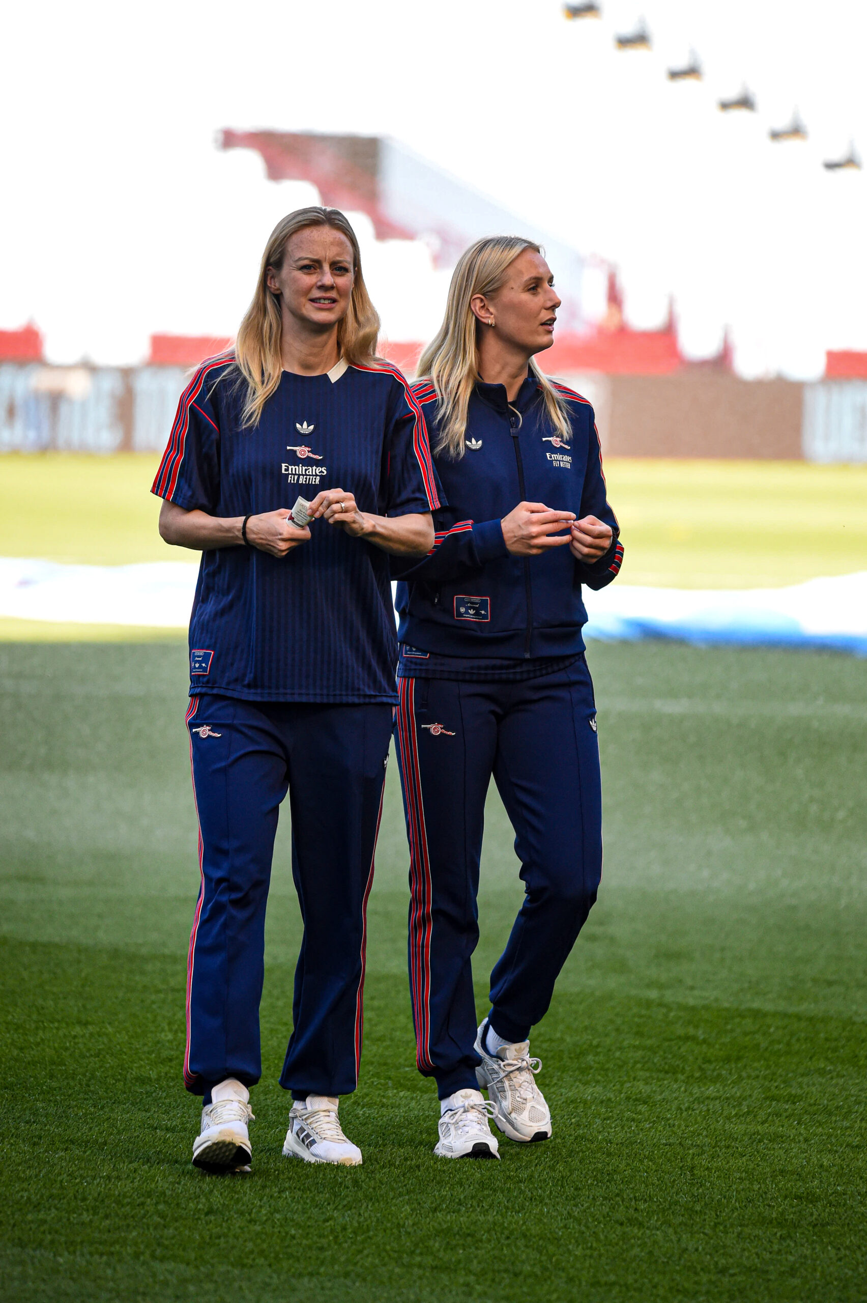 Amanda Ilestedt and Stina Blackstenius wearing matching navy blue Adidas tracksuits with red accents and “Emirates Fly Better” logos walk on a sports field. The woman on the left wears a short-sleeved top, while the woman on the right wears a zip-up jacket. Both have long blonde hair and are holding small items in their hands. The background shows stadium seating and bright daylight.