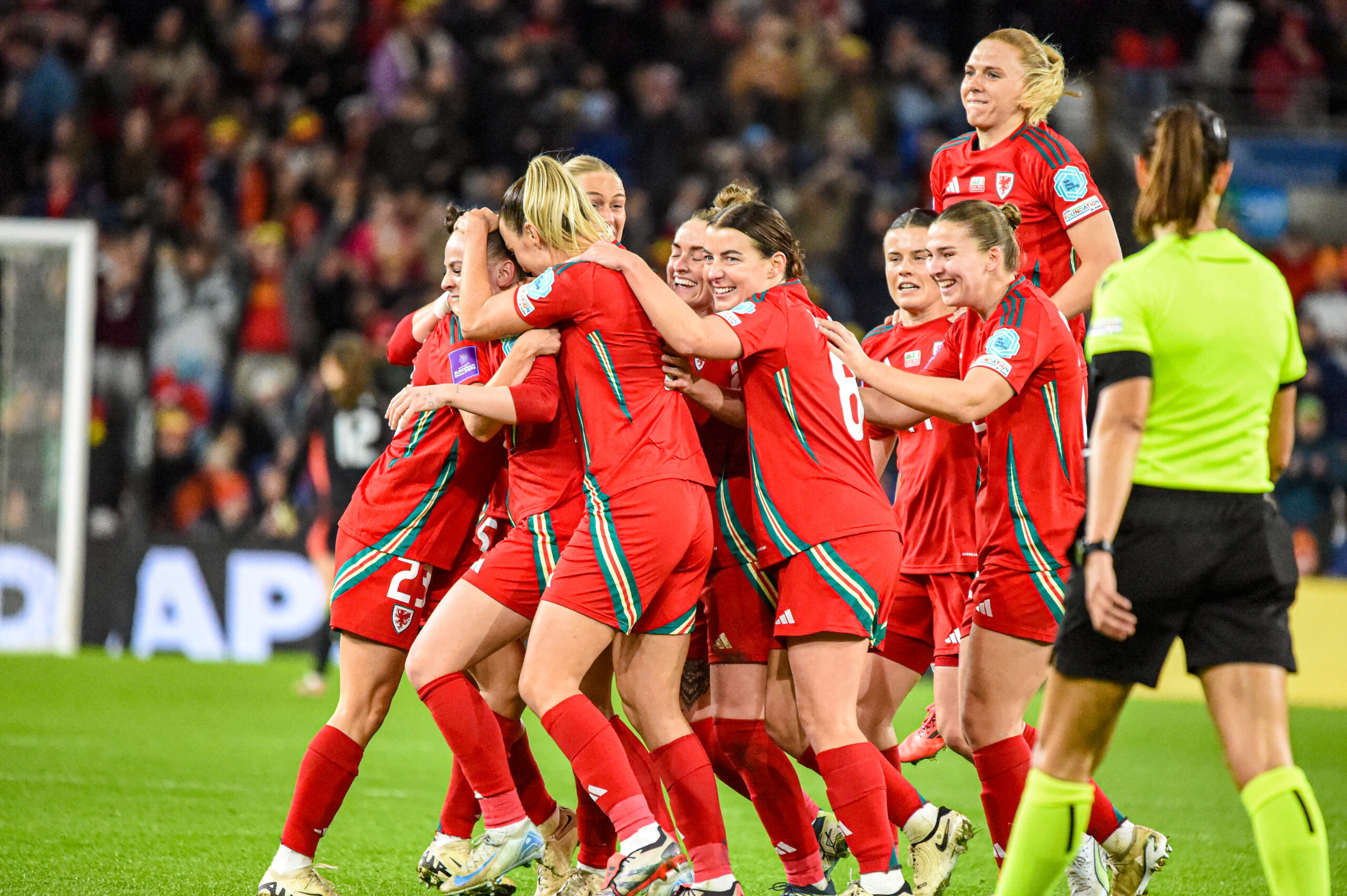A group of Welsh women's national football team players in red uniforms celebrate on the field, hugging and smiling after a successful moment during a match. One player is lifted off the ground in joy. A referee in a neon yellow shirt and black shorts stands nearby. The background shows a cheering crowd in the stadium.