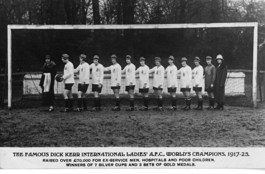 A black and white photograph of a women's football team, the Dick, Kerr Ladies, posing in a line in front of a goalpost on a muddy field. The players are wearing matching uniforms with striped caps, light-coloured long-sleeved jerseys, dark shorts, and striped socks. One woman on the far left and another on the far right are wearing long, dark coats and hats. A text caption below the image reads "THE FAMOUS DICK KERR INTERNATIONAL LADIES' A.F.C., WORLD'S CHAMPIONS, 1917-25. RAISED OVER £70,000 FOR EX-SERVICE MEN, HOSPITALS AND POOR CHILDREN. WINNERS OF 7 SILVER CUPS AND 3 SETS OF GOLD MEDALS."