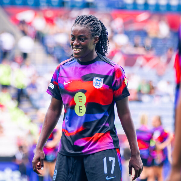 Michelle Agyemang wearing a black and purple patterned jersey with the England national team crest and the number 17 is smiling during a warmup.