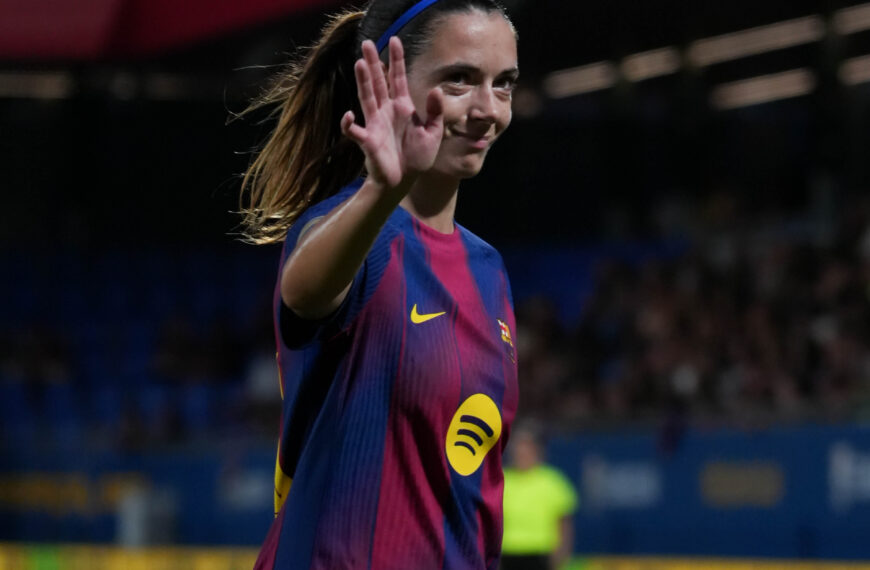 Bonmati is shown from the waist up. She is wearing a dark blue and red striped kit with a yellow swoosh logo and a yellow "Spotify" logo. She has her hair tied back with a blue headband and is smiling, with one hand raised in a waving gesture. The background is a blurry stadium with bright lights and spectators in the stands.