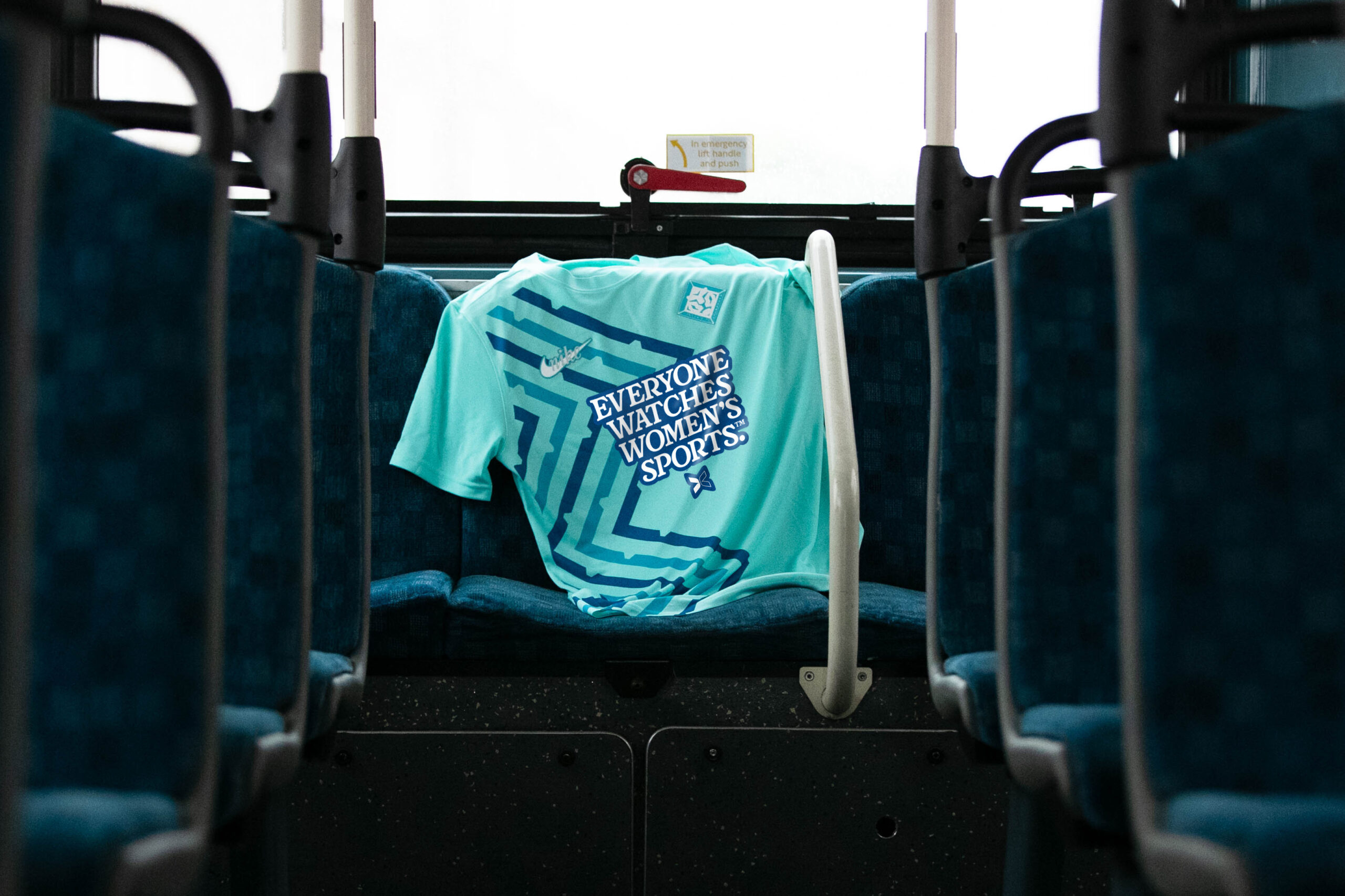 A light blue t-shirt with a darker blue geometric pattern is folded and draped over a seat on a bus. The text "EVERYONE WATCHES WOMEN'S SPORTS" is printed on the front of the shirt.
