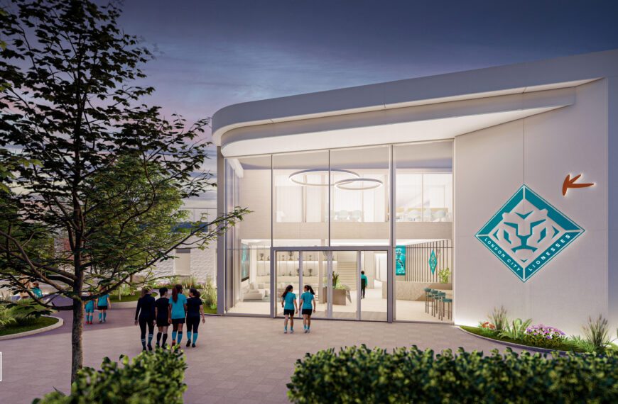 A high-quality architectural rendering of a modern, white building at dusk, which appears to be a sports training or clubhouse facility. The building has a striking entrance with a large, curved glass facade and a prominent London City Lionesses logo in teal and white on the exterior wall to the right. The interior is brightly lit, showcasing a spacious lobby, stairs, and modern seating. Outside, in the foreground, there's paving, landscaping with low bushes, and a mature tree on the left. In the middle ground, a group of people wearing matching teal and black athletic gear are walking toward the entrance. The overall scene is illuminated with soft exterior and interior lighting against the twilight sky.