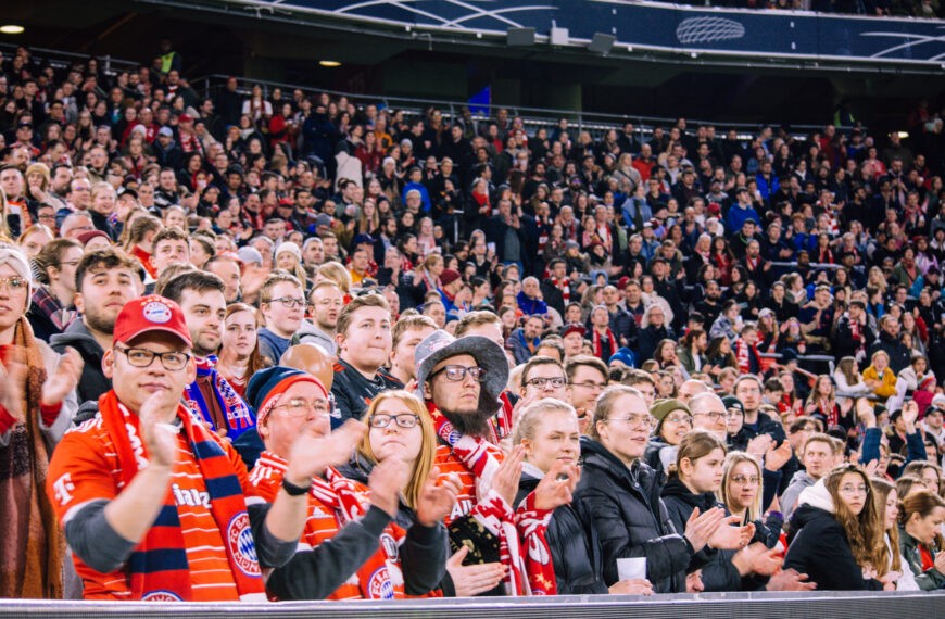A wide-angle photo capturing a large crowd of Bayern Munich fans in the stands of a stadium at night. The foreground shows several people smiling, clapping, and cheering. Many of them are wearing red Bayern Munich jerseys and red and white scarves that feature the club's colors. The background is filled with many more spectators who extend up into the stadium's upper deck, all watching an event on the field that is out of frame. The stadium lighting illuminates the crowd.