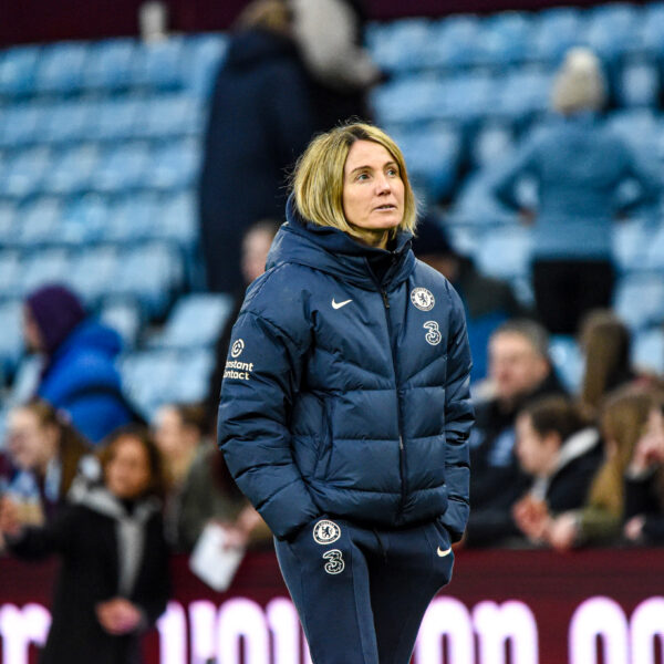 A photograph of Sonia Bompastor, a blonde-haired woman, standing pitch-side at a stadium. She is wearing a dark blue Chelsea FC puffer jacket and matching track pants, looking intently to her right (viewer's left). The background is blurred, showing spectators in the stands, which are primarily blue and claret.