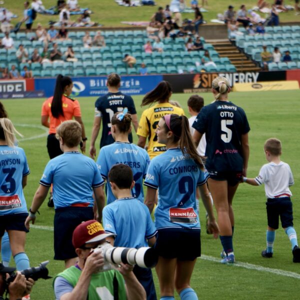 A slightly blurry photograph captures the backs of two teams of football players walking onto a grass pitch at a stadium, accompanied by children and obscured by cameramen in the foreground. The majority of the players are wearing blue jerseys with dark blue shorts, appearing to be one team, with several adult players and a few children in the blue kit, including one young boy holding the hand of a player wearing number 5. Another group of players, possibly the opposing team, are wearing orange/red or yellow/black jerseys, partially visible at the left and center. Many of the adult players have ponytails or their hair tied back. The jerseys have various names and numbers, such as "BRYSON 3" and "CONNOR 9" on the blue team. The players are walking away from the camera toward the far side of the pitch. In the background, there are rows of blue stadium seating, most of which are sparsely occupied by spectators. An advertising banner with "Liberty" and "Maccas" text is visible on the far side of the pitch. The scene suggests a professional women's football match walkout.