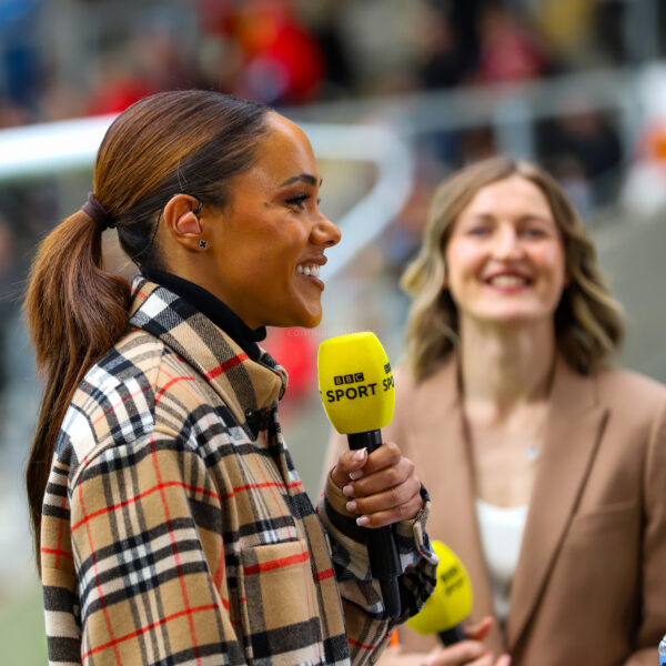 Alex Scott is speaking into a bright yellow BBC Sport microphone while standing at the edge of a stadium pitch. She is shown in profile, smiling, with her hair tied back in a sleek ponytail and wearing a beige, black, and red plaid coat. An in-ear monitor is visible in her right ear. Behind her, slightly out of focus, another presenter with shoulder-length wavy hair and a beige blazer smiles toward the camera. The blurred background shows stadium seating and spectators, suggesting a live sports broadcast environment.