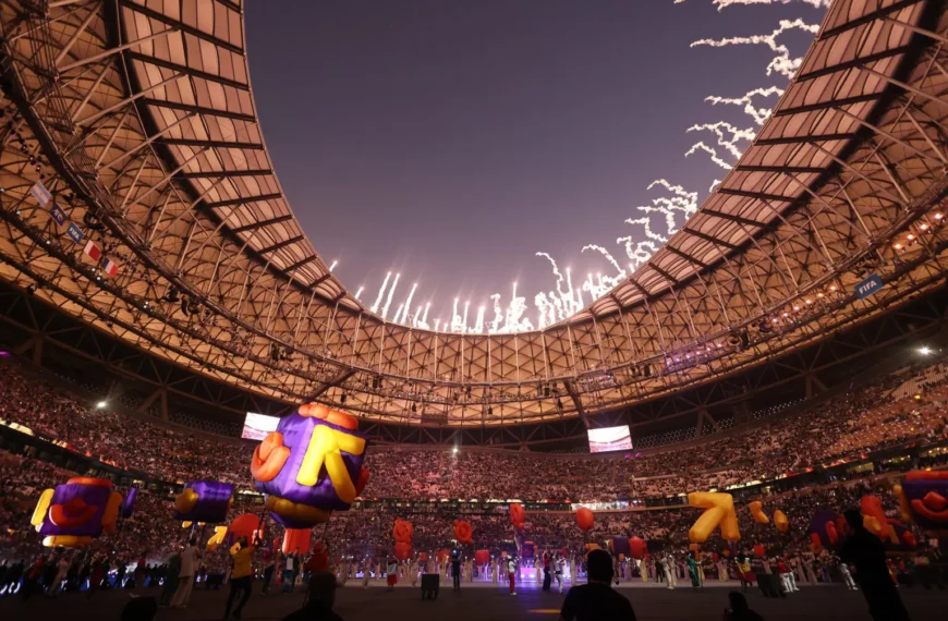 A wide-angle, low-angle shot of a massive, crowded sports stadium during a vibrant opening or closing ceremony at night. The stadium's intricate, golden-hued geometric roof frames a dark sky filled with bright white fireworks and light trails. On the field, large, colourful inflatable characters in purple, orange, and yellow are being moved around by performers. The stands are packed with thousands of spectators, and several large video screens are visible. The overall atmosphere is celebratory and grand.