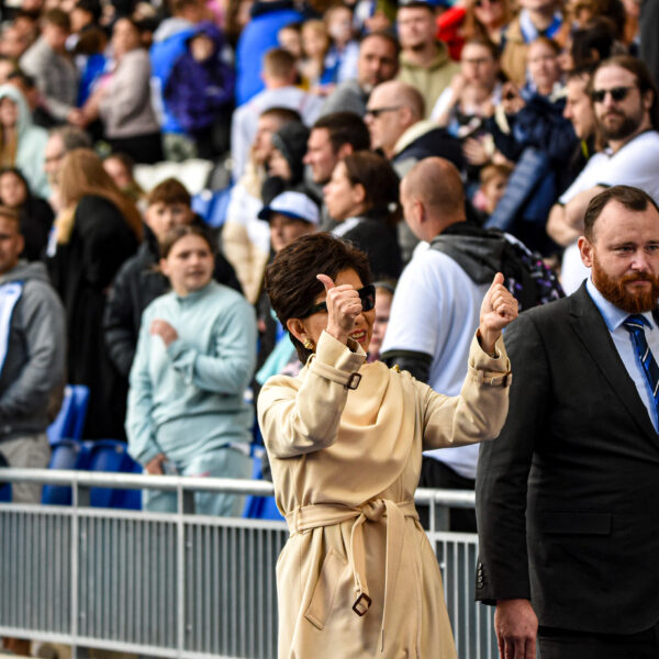 Michele Kang wearing a beige trench coat and sunglasses, smiles brightly while giving two thumbs up. She is the central focus, standing in front of a metal railing at what appears to be a crowded outdoor stadium or sporting event. To her right, a man with a reddish-brown beard, wearing a dark suit and a blue striped tie, walks beside her with a neutral expression. In the background, a large, diverse crowd of spectators fills the stands, some wearing blue and white scarves. The lighting is bright and natural, suggesting an afternoon event.