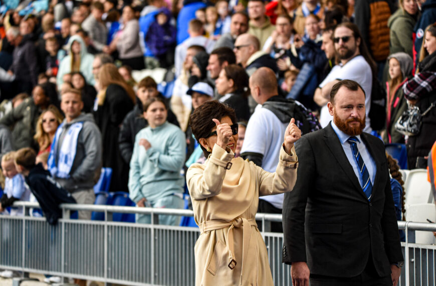 Michele Kang wearing a beige trench coat and sunglasses, smiles brightly while giving two thumbs up. She is the central focus, standing in front of a metal railing at what appears to be a crowded outdoor stadium or sporting event. To her right, a man with a reddish-brown beard, wearing a dark suit and a blue striped tie, walks beside her with a neutral expression. In the background, a large, diverse crowd of spectators fills the stands, some wearing blue and white scarves. The lighting is bright and natural, suggesting an afternoon event.