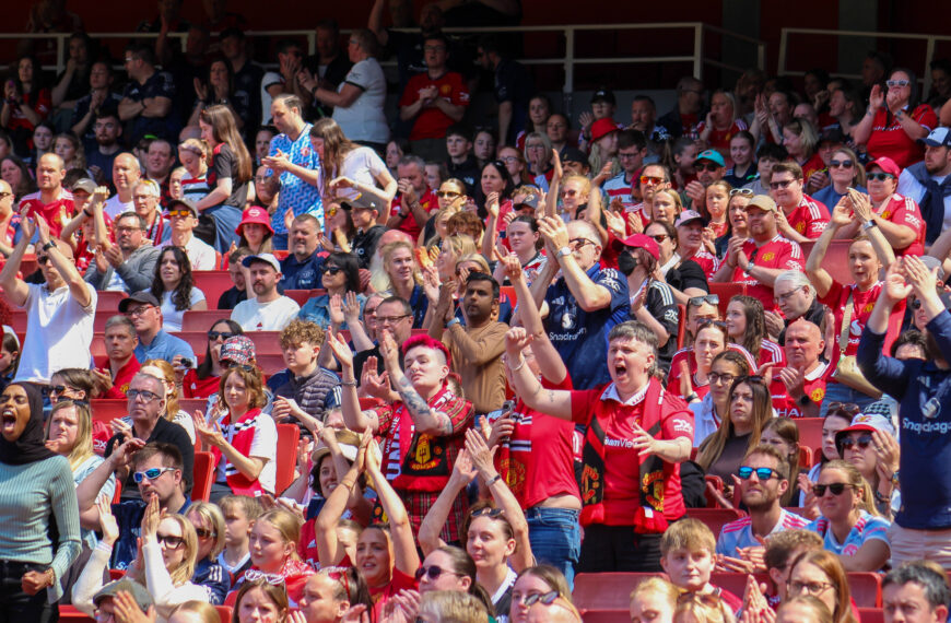 A wide shot of a crowded stadium section filled with Manchester United fans during a match. The crowd is a diverse mix of people of various ages and genders, many wearing the team’s red home shirt, while others wear white or blue away kits and official merchandise. In the foreground and mid ground, fans are shown in various states of excitement: some are cheering with mouths wide open, others are clapping their hands above their heads, and many are wearing sunglasses to shield against the bright sunlight. The red stadium seats are visible between the rows of supporters. The atmosphere appears high-energy and celebratory.