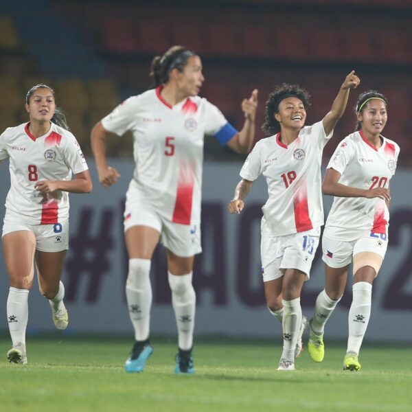 Four female football players in white and red kits celebrate on a green pitch during a match. The player in the centre, wearing number 18, smiles broadly and points upward with one hand, while her teammates run alongside her with expressions of joy and excitement. They are wearing white kits with red accents, white shorts, and tall white socks. In the background, there are blurred stadium seats and a banner with "#WAC2022" visible.