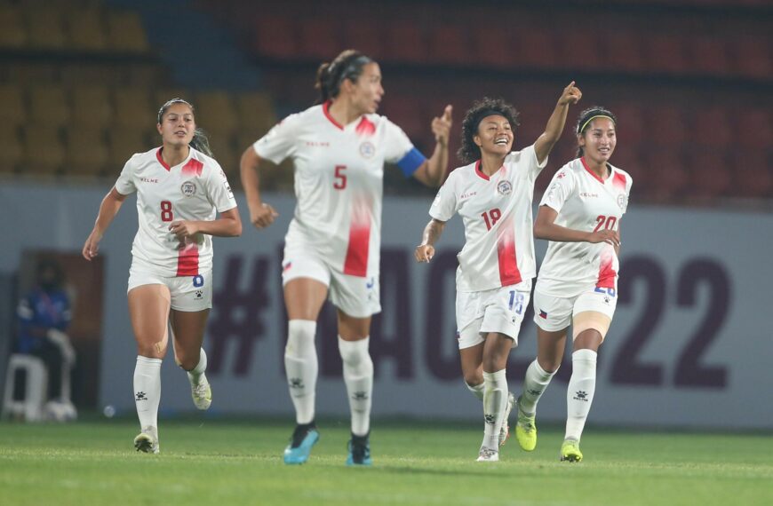 Four female football players in white and red kits celebrate on a green pitch during a match. The player in the centre, wearing number 18, smiles broadly and points upward with one hand, while her teammates run alongside her with expressions of joy and excitement. They are wearing white kits with red accents, white shorts, and tall white socks. In the background, there are blurred stadium seats and a banner with "#WAC2022" visible.