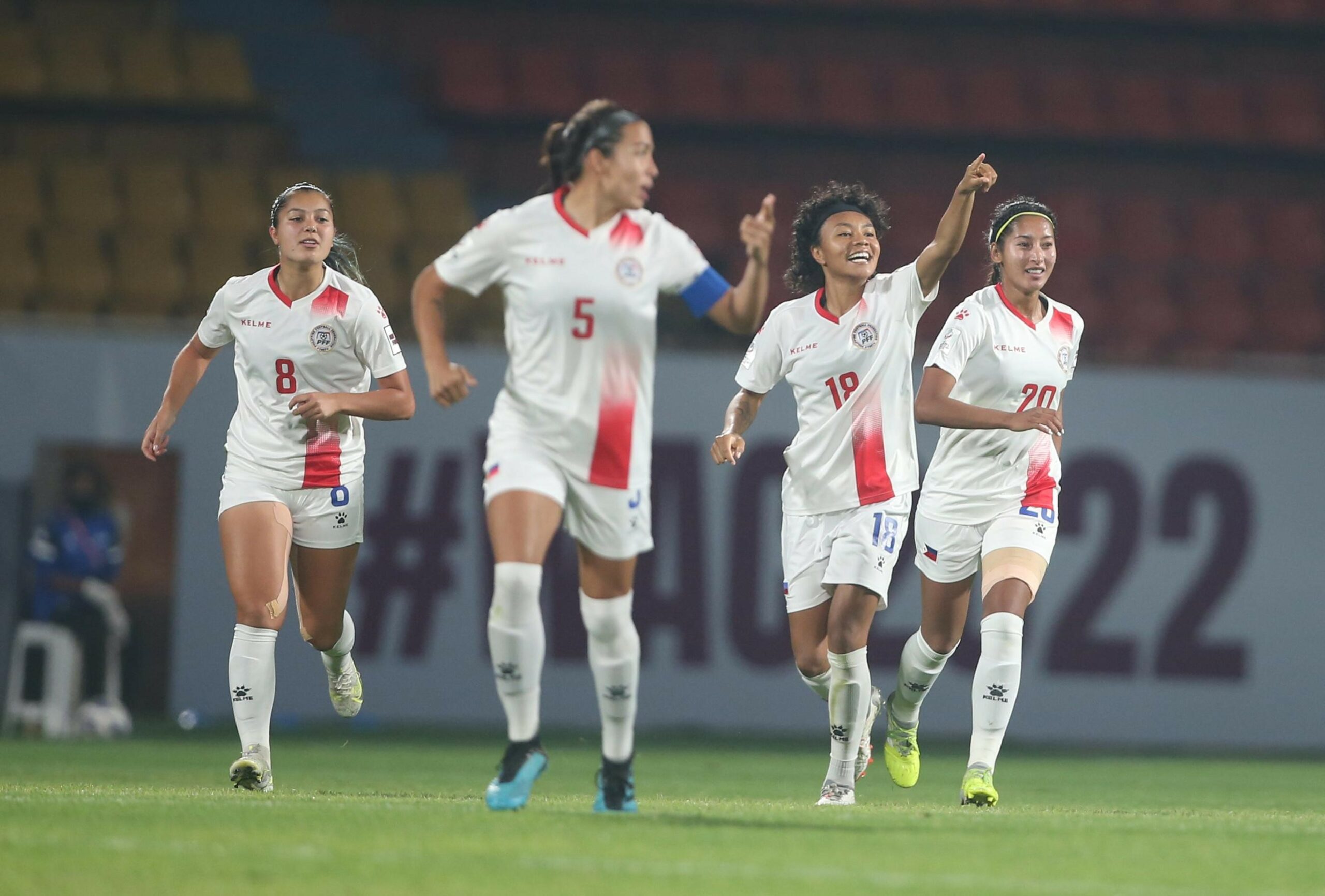Four female football players in white and red kits celebrate on a green pitch during a match. The player in the centre, wearing number 18, smiles broadly and points upward with one hand, while her teammates run alongside her with expressions of joy and excitement. They are wearing white kits with red accents, white shorts, and tall white socks. In the background, there are blurred stadium seats and a banner with "#WAC2022" visible.