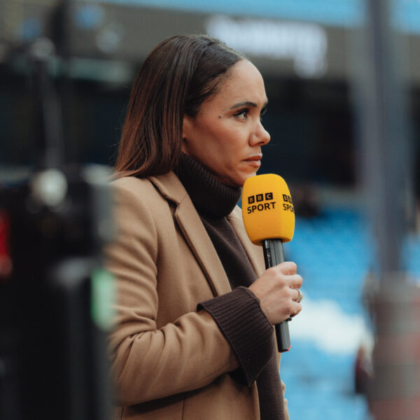 Alex Scott stood holding BBC microphone presenting in brown coat. The picture shows her side profile.