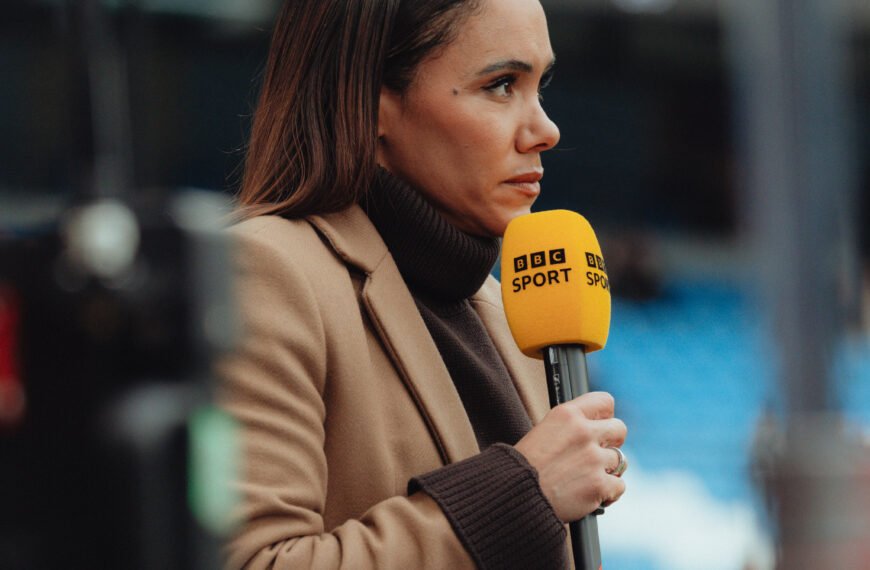 Alex Scott stood holding BBC microphone presenting in brown coat. The picture shows her side profile.