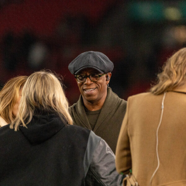 A medium shot captures Ian Wright from the chest up, smiling as he talks with three people whose backs are to the camera. Wright is wearing a dark olive green shawl-collar sweater, a grey flat cap, and round-rimmed glasses. To his right, a person in a tan coat has a thin white earbud wire running down their back. The group is standing in a stadium with a blurred background of red seats and green pitch.