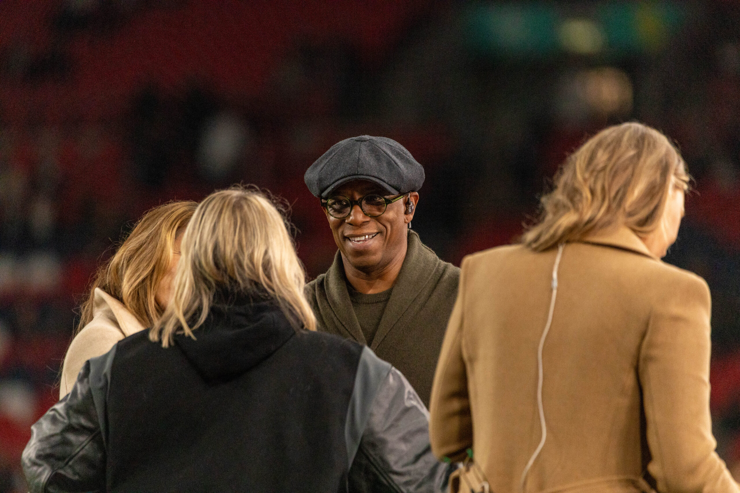A medium shot captures Ian Wright from the chest up, smiling as he talks with three people whose backs are to the camera. Wright is wearing a dark olive green shawl-collar sweater, a grey flat cap, and round-rimmed glasses. To his right, a person in a tan coat has a thin white earbud wire running down their back. The group is standing in a stadium with a blurred background of red seats and green pitch.