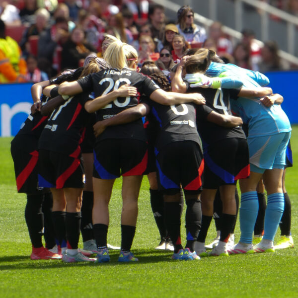 A group of female players from Olympique Lyonnais Féminin in a huddle on a sunny grass pitch. Most players wear black kits with red and blue accents, while the goalkeeper is in a light blue kit. In the background, a crowd of spectators sits in stadium seats.