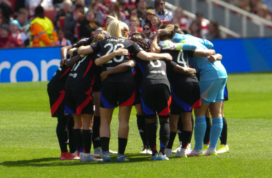 A group of female players from Olympique Lyonnais Féminin in a huddle on a sunny grass pitch. Most players wear black kits with red and blue accents, while the goalkeeper is in a light blue kit. In the background, a crowd of spectators sits in stadium seats.