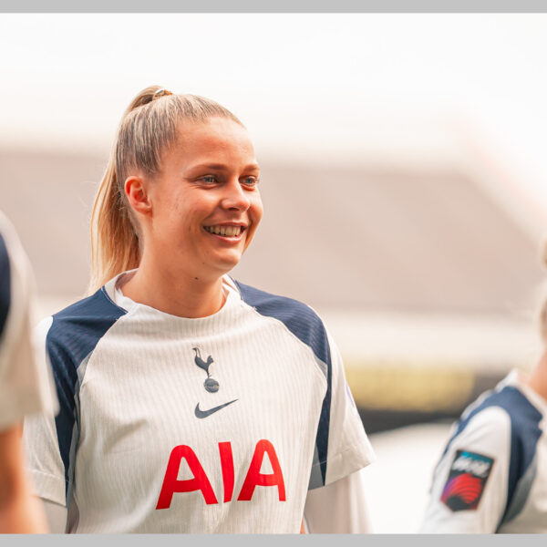 A medium shot of Olivia Holdt, female football player for Tottenham Hotspur. She has blonde hair tied back in a high ponytail and is wearing the team's white and navy blue kit featuring the AIA sponsor logo and the Nike swoosh. The background is a brightly lit, slightly blurred outdoor stadium setting.