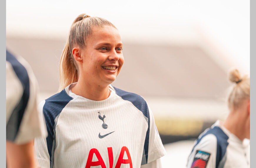 A medium shot of Olivia Holdt, female football player for Tottenham Hotspur. She has blonde hair tied back in a high ponytail and is wearing the team's white and navy blue kit featuring the AIA sponsor logo and the Nike swoosh. The background is a brightly lit, slightly blurred outdoor stadium setting.