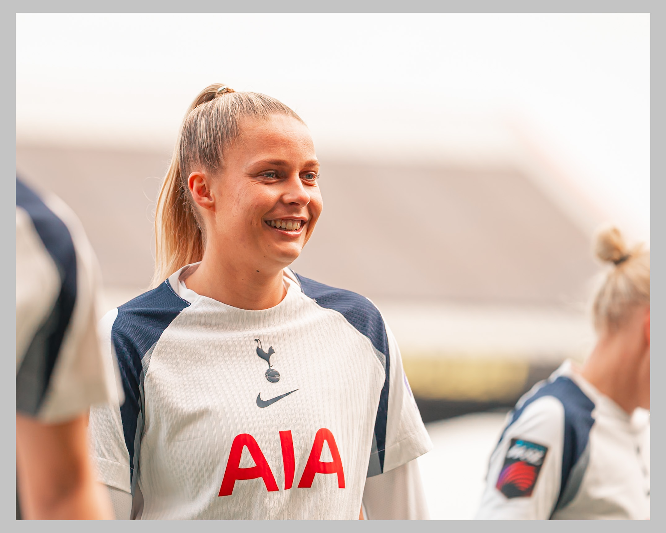 A medium shot of Olivia Holdt, female football player for Tottenham Hotspur. She has blonde hair tied back in a high ponytail and is wearing the team's white and navy blue kit featuring the AIA sponsor logo and the Nike swoosh. The background is a brightly lit, slightly blurred outdoor stadium setting.
