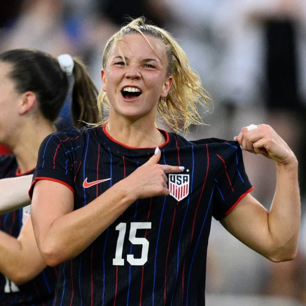 A close-up shot shows Claire Hutton for the United States women's national team celebrating. She is wearing a black football kit with thin red and blue vertical stripes, the number 15, and the USA crest. With a joyous, open-mouthed expression, she pulls at the collar of her jersey and points directly at the team crest with her thumb. Her blonde hair is pulled back in a ponytail, with several damp strands framing her face. Another player is partially visible in the blurred background.