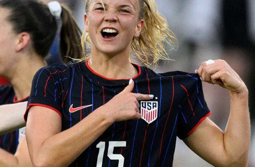 A close-up shot shows Claire Hutton for the United States women's national team celebrating. She is wearing a black football kit with thin red and blue vertical stripes, the number 15, and the USA crest. With a joyous, open-mouthed expression, she pulls at the collar of her jersey and points directly at the team crest with her thumb. Her blonde hair is pulled back in a ponytail, with several damp strands framing her face. Another player is partially visible in the blurred background.