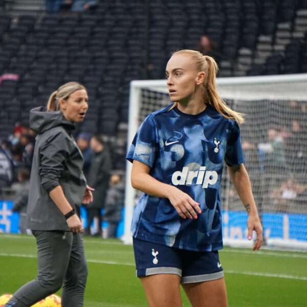Molly Bartrip stands on a green pitch wearing a blue patterned training top with "drip" on the front and dark blue shorts featuring the Tottenham Hotspur crest. In the background, a woman in a grey hooded jacket looks toward the player, while a white goal frame and empty stadium seating are visible behind them. Cones and balls sit on the grass in the foreground.