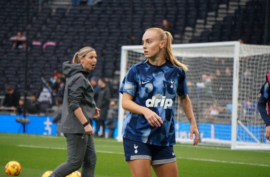 Molly Bartrip stands on a green pitch wearing a blue patterned training top with "drip" on the front and dark blue shorts featuring the Tottenham Hotspur crest. In the background, a woman in a grey hooded jacket looks toward the player, while a white goal frame and empty stadium seating are visible behind them. Cones and balls sit on the grass in the foreground.
