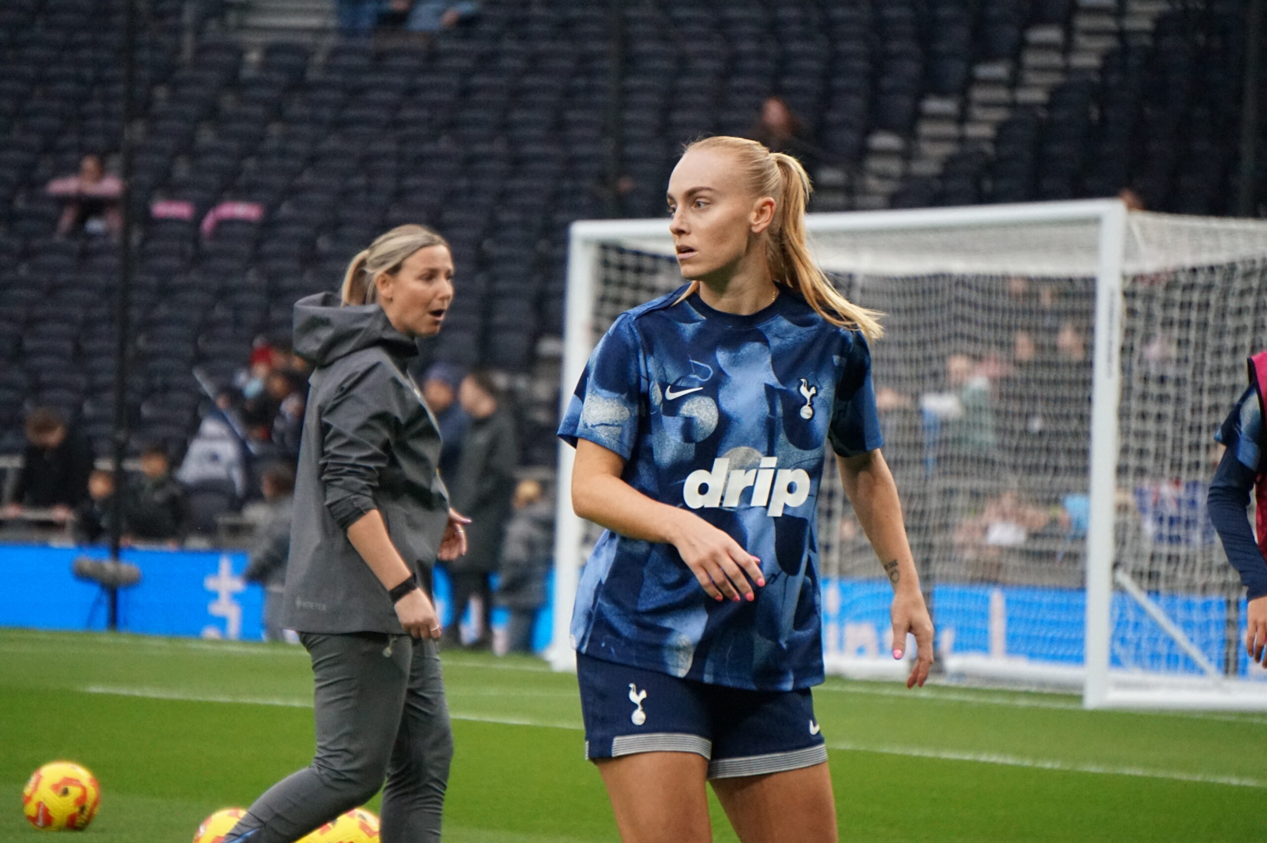 Molly Bartrip stands on a green pitch wearing a blue patterned training top with "drip" on the front and dark blue shorts featuring the Tottenham Hotspur crest. In the background, a woman in a grey hooded jacket looks toward the player, while a white goal frame and empty stadium seating are visible behind them. Cones and balls sit on the grass in the foreground.