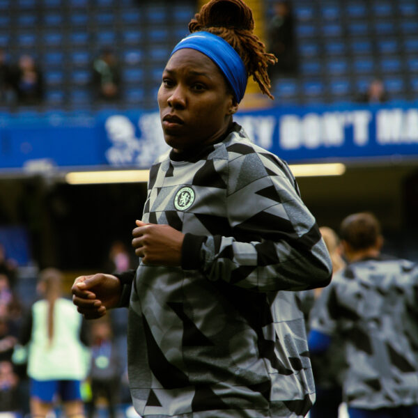 A close-up, medium shot of a Kadeisha Buchanan during a warm-up. She is wearing a grey and black patterned Chelsea F.C. training top and a bright blue headband that holds back her dreadlocks, which are styled in a high bun. She has a focused expression as she jogs on the pitch. The background shows the blurred blue seats of a stadium and other players in the distance.