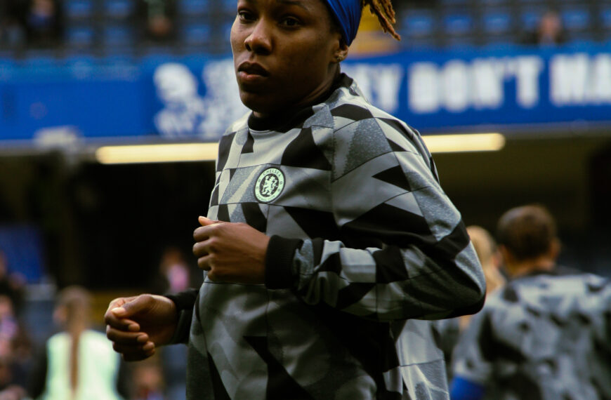 A close-up, medium shot of a Kadeisha Buchanan during a warm-up. She is wearing a grey and black patterned Chelsea F.C. training top and a bright blue headband that holds back her dreadlocks, which are styled in a high bun. She has a focused expression as she jogs on the pitch. The background shows the blurred blue seats of a stadium and other players in the distance.