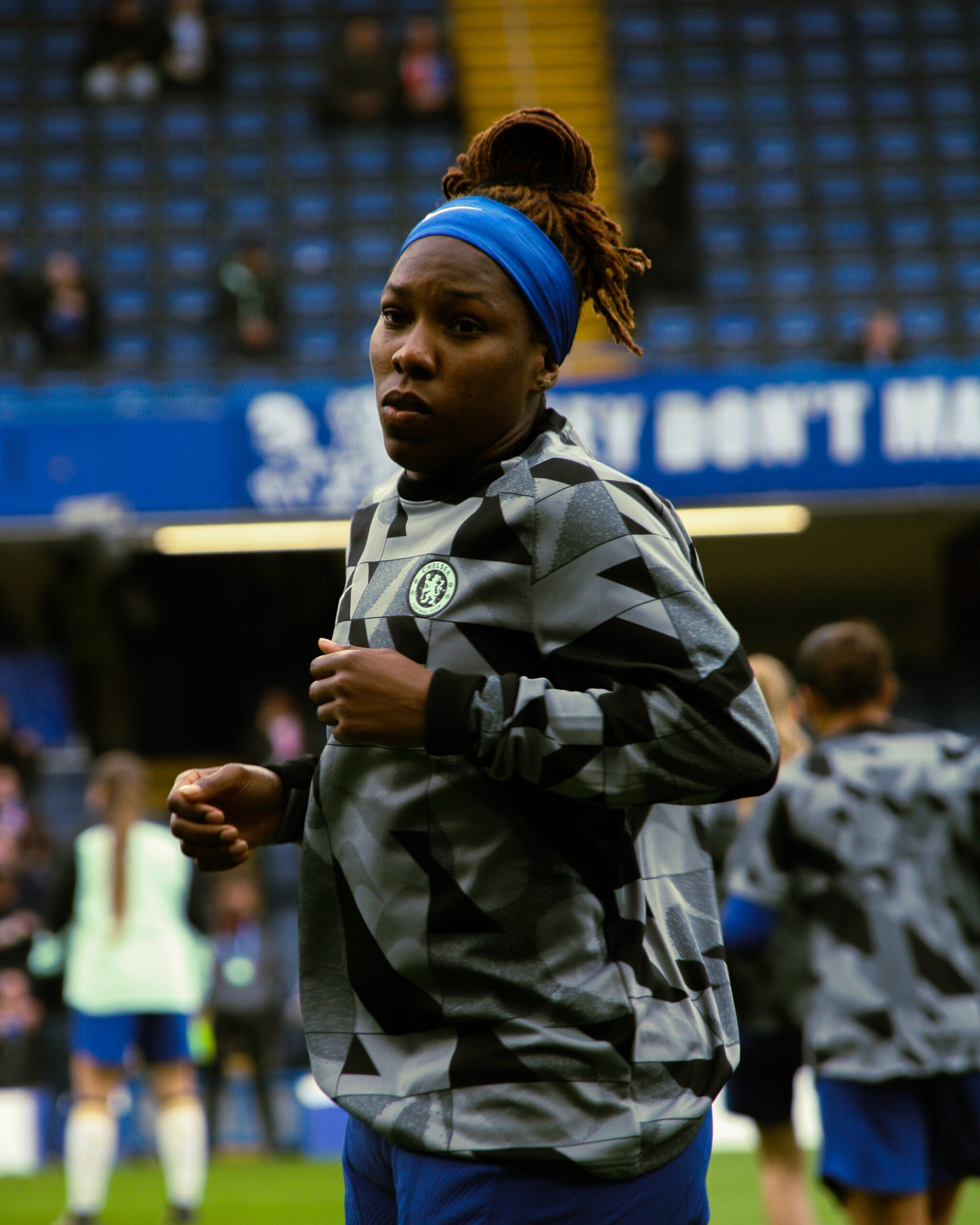 A close-up, medium shot of a Kadeisha Buchanan during a warm-up. She is wearing a grey and black patterned Chelsea F.C. training top and a bright blue headband that holds back her dreadlocks, which are styled in a high bun. She has a focused expression as she jogs on the pitch. The background shows the blurred blue seats of a stadium and other players in the distance.