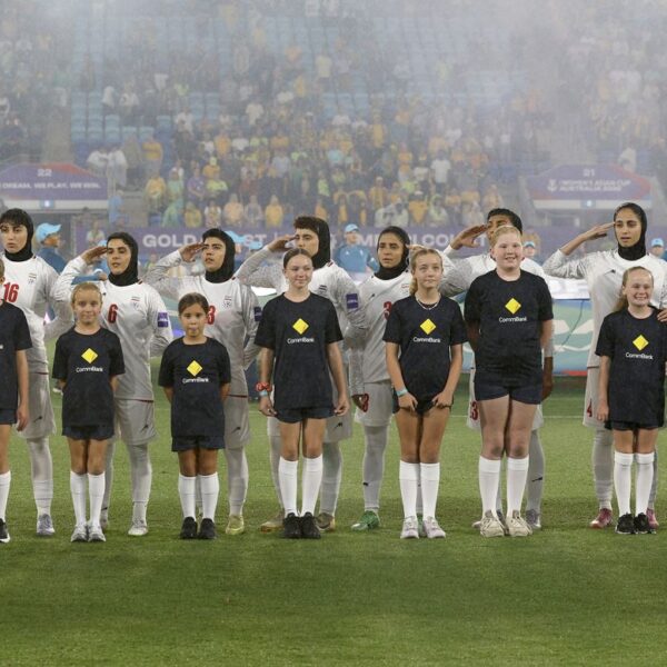 In a wide, eye-level shot, eleven members of the Iranian women's national football team stand in a line on a green pitch, wearing white kits and hijabs. Most of the players have their right hands raised to their foreheads in a salute, their expressions solemn. In front of them, eleven young mascots in dark blue t-shirts with a yellow diamond logo and matching shorts stand in a row. Some of the children are smiling and holding hands. In the background, a large, blurred crowd of spectators fills the stadium seats, with faint banners and signs visible through a light mist or fog. A blue and white flag is partially visible behind the players.
