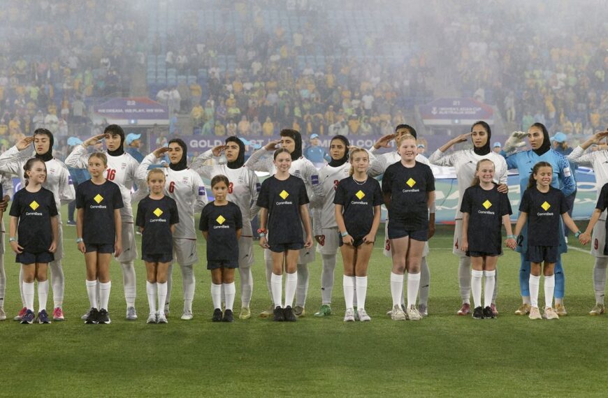 In a wide, eye-level shot, eleven members of the Iranian women's national football team stand in a line on a green pitch, wearing white kits and hijabs. Most of the players have their right hands raised to their foreheads in a salute, their expressions solemn. In front of them, eleven young mascots in dark blue t-shirts with a yellow diamond logo and matching shorts stand in a row. Some of the children are smiling and holding hands. In the background, a large, blurred crowd of spectators fills the stadium seats, with faint banners and signs visible through a light mist or fog. A blue and white flag is partially visible behind the players.