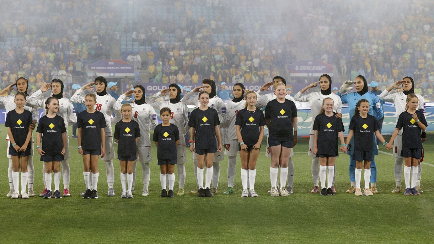 In a wide, eye-level shot, eleven members of the Iranian women's national football team stand in a line on a green pitch, wearing white kits and hijabs. Most of the players have their right hands raised to their foreheads in a salute, their expressions solemn. In front of them, eleven young mascots in dark blue t-shirts with a yellow diamond logo and matching shorts stand in a row. Some of the children are smiling and holding hands. In the background, a large, blurred crowd of spectators fills the stadium seats, with faint banners and signs visible through a light mist or fog. A blue and white flag is partially visible behind the players.