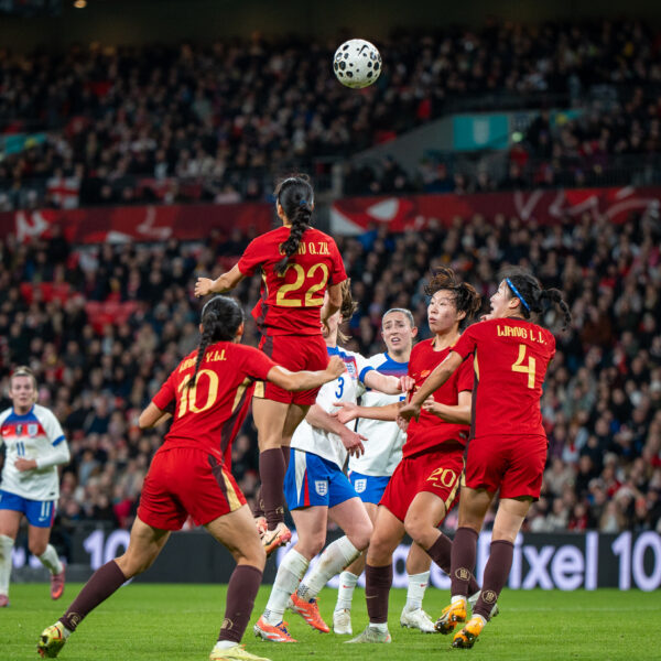 Action shot from a women's international football match between England (the Lionesses) and China. In the foreground, five players are tightly clustered as they contest a high ball. A Chinese player wearing a red and gold jersey with the number 22 is leaping high into the air to head a black-and-white patterned football. Surrounding her are two teammates in red (numbers 10 and 4) and two England players in white and blue kits. The stadium is packed with spectators in the background, under bright floodlights, with a digital advertising board for the "Google Pixel 10" visible behind the players. The atmosphere appears intense and competitive.