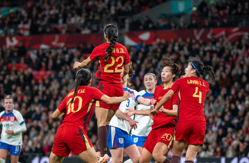 Action shot from a women's international football match between England (the Lionesses) and China. In the foreground, five players are tightly clustered as they contest a high ball. A Chinese player wearing a red and gold jersey with the number 22 is leaping high into the air to head a black-and-white patterned football. Surrounding her are two teammates in red (numbers 10 and 4) and two England players in white and blue kits. The stadium is packed with spectators in the background, under bright floodlights, with a digital advertising board for the "Google Pixel 10" visible behind the players. The atmosphere appears intense and competitive.
