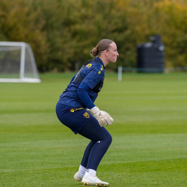 A side profile of Maddison Millington-Stanbury, goalkeeper, during a training session. The player is in a focused, crouched "ready position" on a green grass field. She is wearing a navy blue long-sleeved training top, navy leggings, white goalkeeper gloves, and white boots. Her hair is pulled back into a neat bun. In the background, there is a white goal and a line of green and yellow trees under an overcast sky.